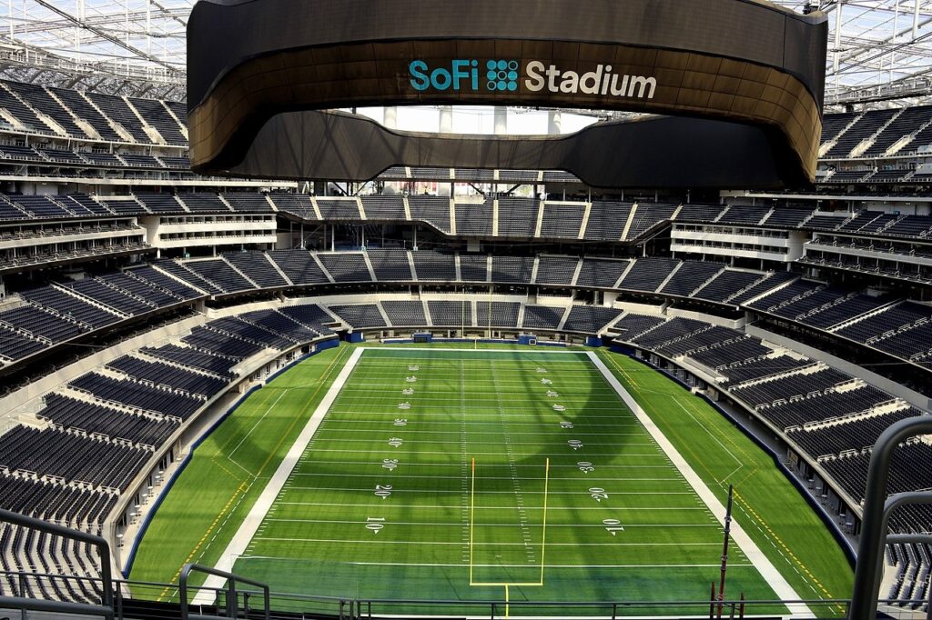 Interior view of SoFi Stadium in Los Angeles showing the football field, multi tier seating, and the large suspended oval video scoreboard above the field.